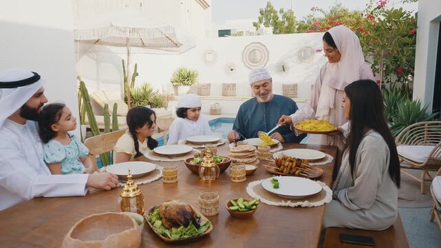 Arab family wearing traditional emirati clothing having lunch at home in the backyard garden - Middle-eastern family bonding and having fun on a family reunion dinner
