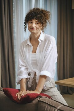Cute woman with curly hair dressed in a white shirt and beige pants stands by a couch in a cozy living room, lightly resting her hands on a red pillow. Soft afternoon light filters through the curtains