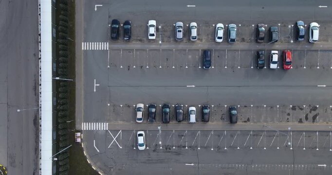 Cinematic top-down aerial flyover shot of a half-empty asphalt parking lot with parked cars and geometric road markings