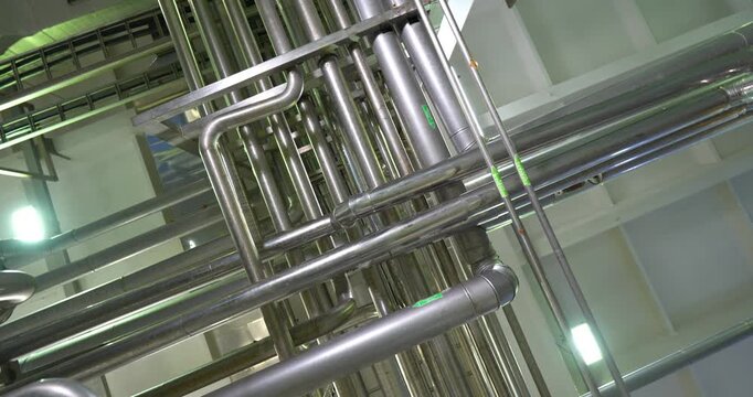 Dynamic low angle shot with a rotating camera looking up at a complex network of stainless steel pipes on the ceiling of an industrial plant