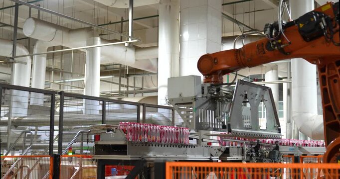 Steady shot of an automated orange industrial robotic arm palletizing packaged beverages behind a safety fence in a modern manufacturing plant