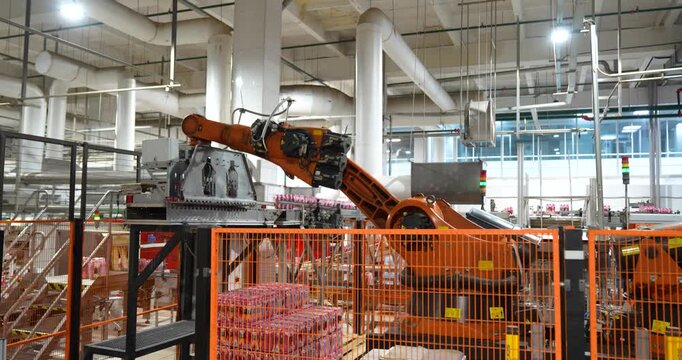 Steady shot of an automated orange industrial robotic arm palletizing packaged beverages behind a safety fence in a modern manufacturing plant