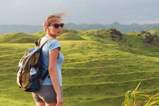 Woman with backpack standing on green grass hill with hair blowing in wind and looking aside. Outdoor adventure concept.