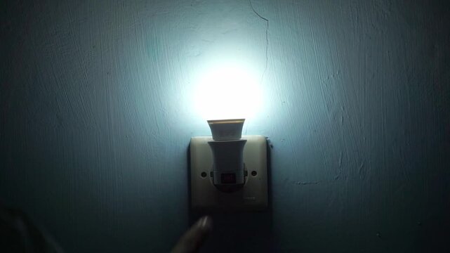 Hand turning on wall light switch, electricity and home lighting concept with glowing LED bulb in dark room interior.