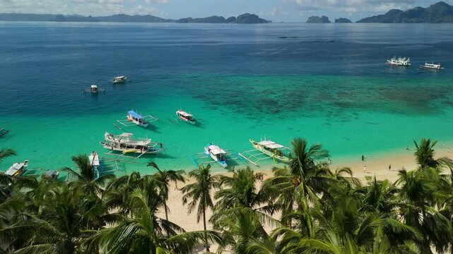 Dozens of outrigger bangka boats moored at Seven Commandos Beach El Nido seen through swaying palm canopy over turquoise reef and sandy shore