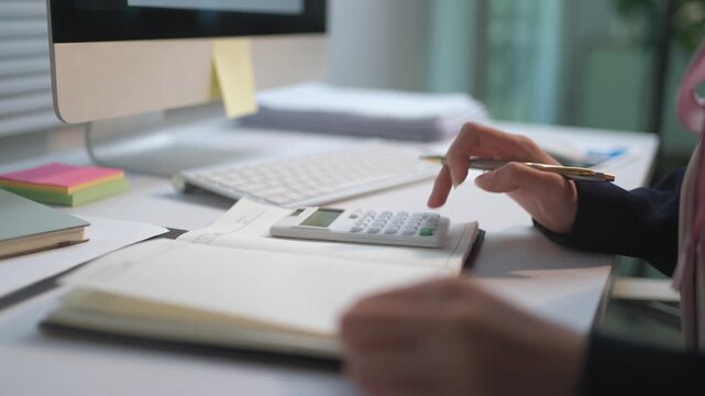 Close up of a female accountant working in an office, using a calculator and writing financial data on her agenda with a pen