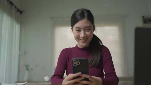 Cheerful young asian woman sitting at desk, happily scrolling and typing on her smartphone. Modern lifestyle and digital communication concept