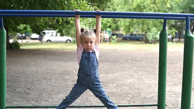 Cute little girl holding onto horizontal bar in park playground, dressed in casual clothes. Girl demonstrates strength and playful energy during outdoor fitness activity in natural setting.