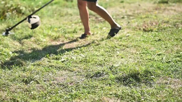 Person using string trimmer to cut tall grass and weeds in sunny garden, closeup of hands operating tool, outdoor landscaping activity, green field and flowers visible, gardening, lawn care concept.