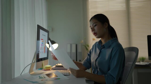 Focused young asian businesswoman comparing documents with business charts on computer screen while working overtime in a dark office at night