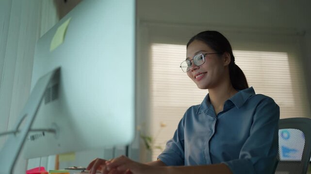 Young professional Asian woman with glasses typing on her computer keyboard. Focused employee working overtime in a modern office at night