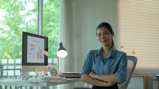 Young asian business professional working on her computer, then turning to smile confidently at the camera with her arms crossed