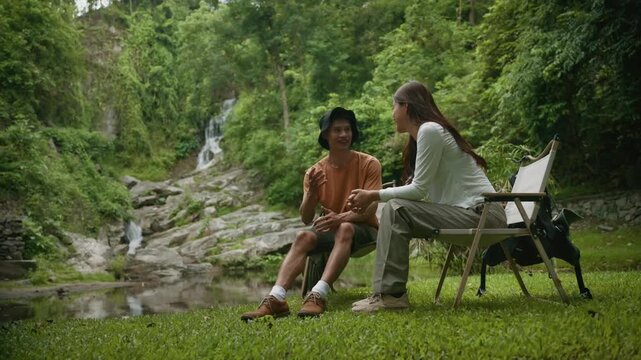 Side view of a beautiful young couple sitting on camping chairs, talking animatedly in a lush green forest with a waterfall in the background
