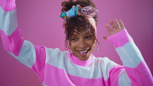 Happy young woman with curly hair and a colorful headband dancing energetically against a vibrant pink background, smiling and gesturing joyfully to the rhythm of music in a studio setting