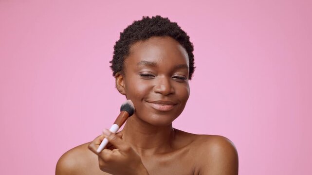Smiling Young Black Woman Applying Blush With Makeup Brush On Cheek, Standing In Front Of Mirror At Home, Taking Care Of Skin. Beauty Routine Concept