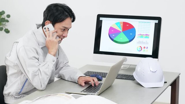 オフィスでPCを見ながらスマホで通話する男性作業員