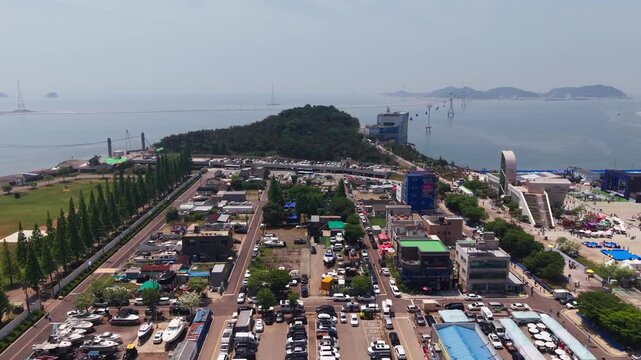 Aerial view of Jeongok Port parking lots filled with cars and trailered yachts, tree-lined boulevard and Daebudo cable car crossing hazy sea past forested headland in South Korea