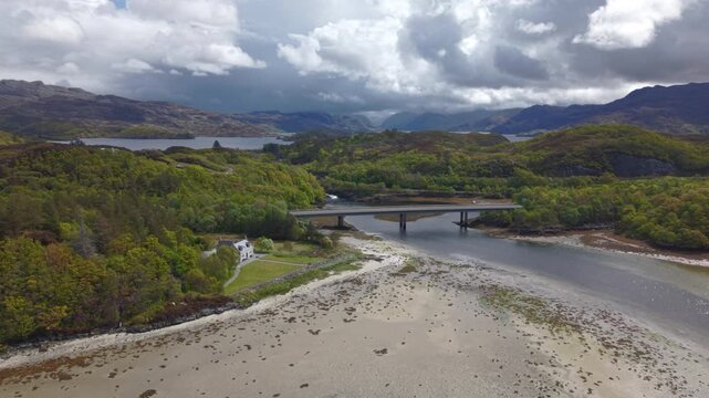 Panning drone shot showcasing the Morar Bridge, tidal river channel, and coastal sandbanks with the sweeping peaks of the Scottish Highlands in the background.
