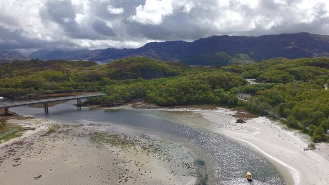 High-angle static drone shot capturing the Morar Bridge, tidal river channel, and the white sands of Morar Beach with the rugged Scottish Highlands in the background.
