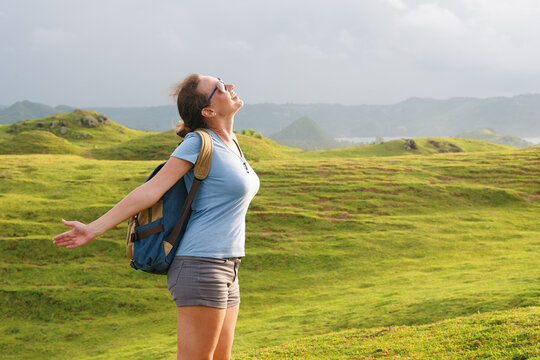 Carefree woman with backpack standing on green grass hill with arms wide open, enjoying wind. Freedom and hiking lifestyle concept.