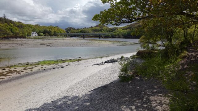 Drone reveal shot rising up past overhanging trees on Morar Beach to uncover the wide tidal estuary and Morar Bridgein the background.