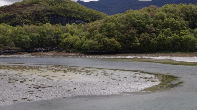 Telephoto drone flight climbing up and pushing forward over the wooded shores of Morar Beach to reveal the dramatic, rugged peaks of the Scottish Highlands behind.