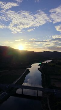 puente sobre rio con solHigh-altitude drone shot tracks steadily forward over a calm river and bridge, revealing the bright sun setting behind dark hills under a blue and cloudy sky.