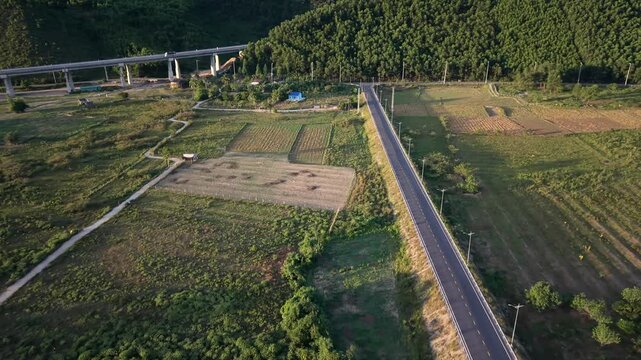 High-altitude drone shot pans and tracks sideways over green countryside fields, revealing a long asphalt road, farming patches, and a massive concrete highway bridge structure stretching across the m