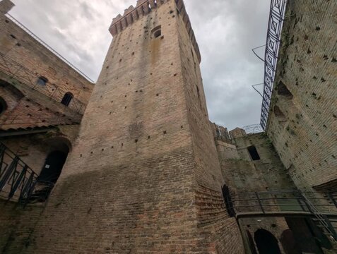 Torre medievale in mattoni ripresa dal basso, con merlature imponenti e atmosfera drammatica sotto un cielo grigio, ideale per contenuti storici e architettonici