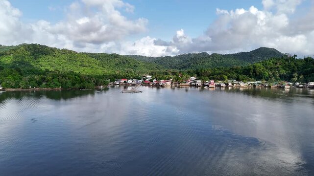 Aerial drone view of a waterfront fishing village with colorful stilted houses along a calm bay backed by densely forested green hills under partly cloudy skies, Taytay, Palawan, Philippines.