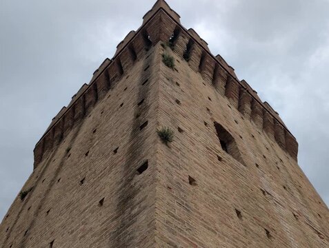 Torre medievale in mattoni ripresa dal basso, con merlature imponenti e atmosfera drammatica sotto un cielo grigio, ideale per contenuti storici e architettonici