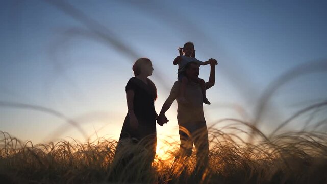 A woman and a man hold hands while a girl sits on the man's shoulders. The scene includes tall grass and a sunset in the background, creating a serene atmosphere.