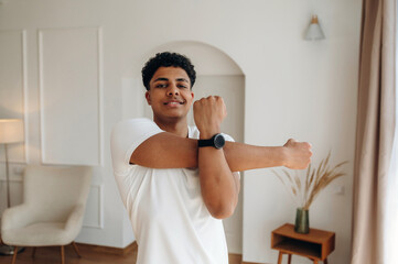 Young African American man in white t-shirt stretches arms inside a modern living room with minimalist decor and natural light streaming through the window © standret