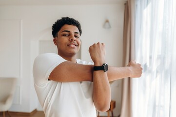 Young male stretching his arms while wearing a white t-shirt and a black wristwatch in a bright, modern indoor living space with natural light © standret