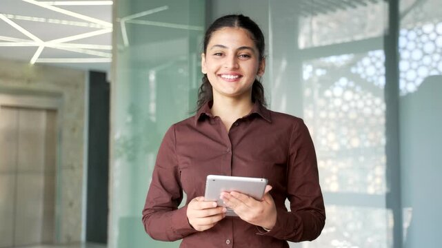 Portrait of smiling businesswoman using a digital tablet and looking at camera at workplace in modern business office. Young female employee reading message, chatting online with a client or browsing.