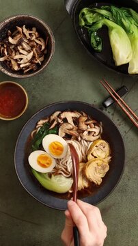 Top view vertical video of hand sprinkling sesame seeds over ramen soup. Final garnish stage of Asian noodle dish preparation on dark flat lay background.