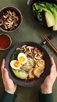 Top view vertical video of hands rotating a finished ramen bowl for presentation. Traditional Asian noodle soup with chicken, egg, pak choi, shiitake mushrooms and garnishes on dark background.