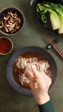Top view vertical video of adding ramen noodles into hot broth. Asian soup assembly process filmed as flat lay on dark background with traditional ingredients and chopsticks.