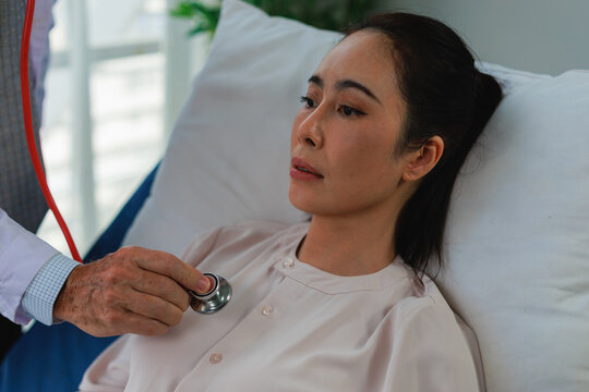 Doctor performing a health check on an Asian female patient with a stethoscope, carefully listening to her heart and lungs during a medical examination in a hospital room