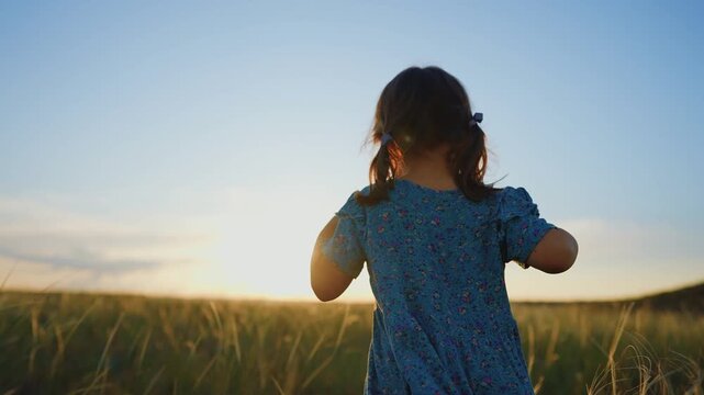 A girl stands in a field, looking towards the sunset. The girl s dress sways gently in the breeze as the sun sets over the grassy landscape.