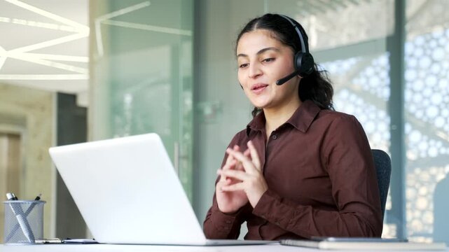 Confident businesswoman in a wireless headset talking on video call using laptop sitting at workplace in office. Young woman manager has business meeting, speaks remotely at an online conference.