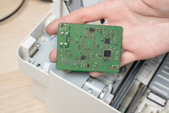 Hand Holding Green Circuit Board Inside Disassembled Printer During Electronics Repair And Maintenance