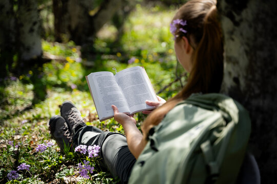 A woman is enjoying a book while relaxing in the woods, surrounded by spring green. Outdoor reading is a concept of mindfulness and a slow life. Back view.
