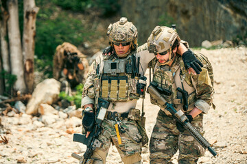 Fototapeta premium Two soldiers in camouflage on rocky terrain; one supports an injured teammate as they move through the field, rifles and tactical gear visible during an outdoor operation.