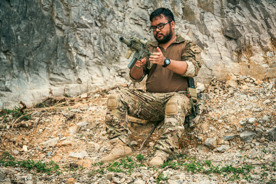 A man in camouflage tactical gear sits by a rocky cliff, inspecting a rifle during an outdoor training break.