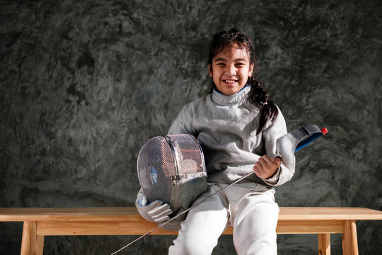 Smiling young fencer in full gear sits on a bench, holding an epee and fencing mask against a dark studio background.