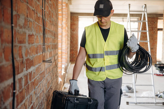 Male construction worker in safety vest carrying electrical cables and toolbox inside a building under renovation with brick walls and a ladder in the background