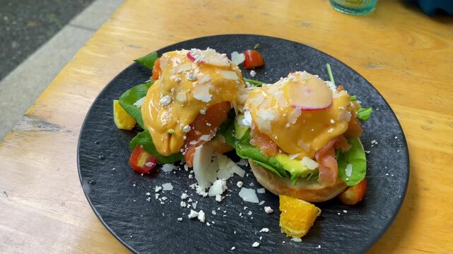 Gourmet eggs Benedict topped with smoked salmon, avocado, poached eggs, and creamy hollandaise sauce. Served on a black plate with fresh greens, parmesan shavings, and citrus garnish in a caf&eacute; setting