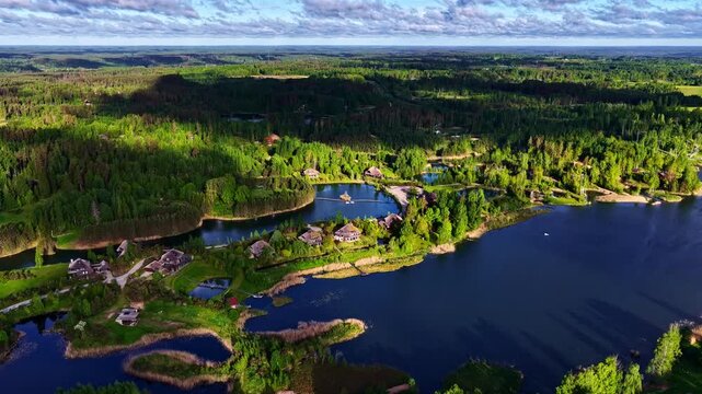 Spectacular wide drone panorama over an exclusive lakeside village community. Features premium thatch-roof homes built along scenic peninsulas surrounded by deep blue waters and dense green forests.