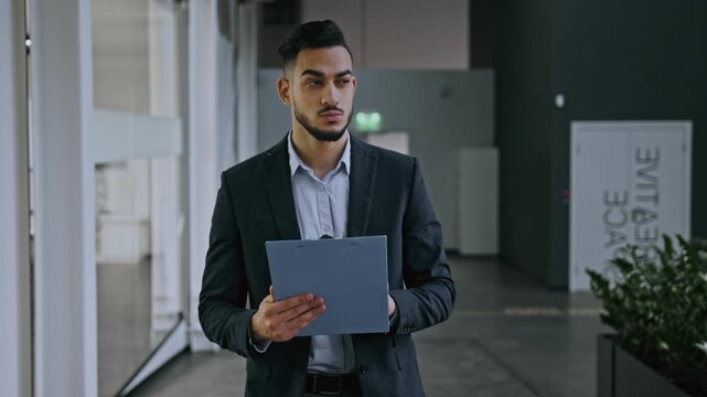 Focused Young Asian Man Dressed In Formal Attire Holding Clipboard While Standing In Office Setting. Professional Male Reviewing Documents In Modern Business Environment.